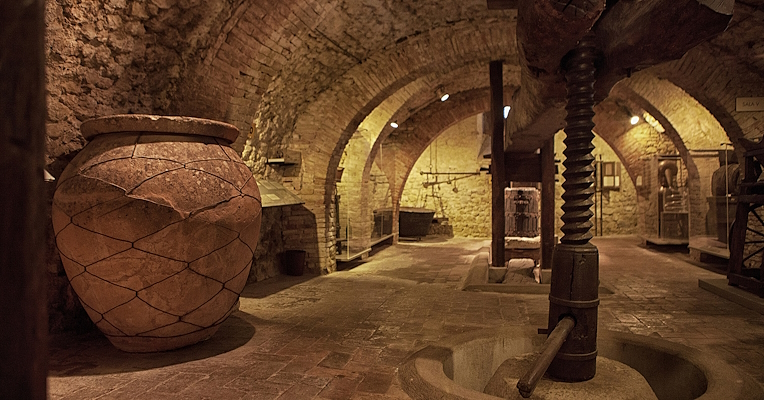 Stone interior of a historic olive press with a wooden press and an amphora, lit by warm lights