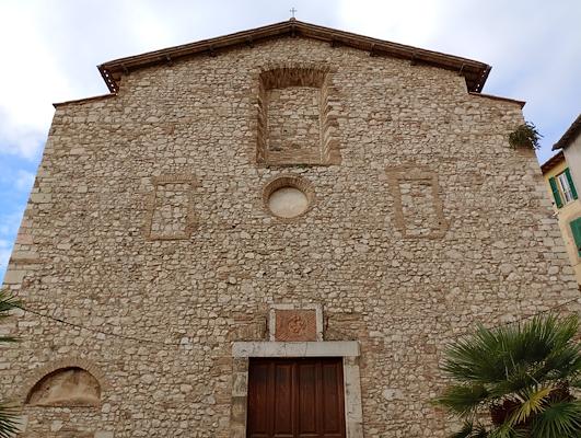 Stone façade of an ancient church with a wooden portal and walled-up windows, surrounded by buildings and plants