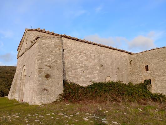 Ancient stone building, surrounded by vegetation and hills, under a blue sky with scattered clouds.