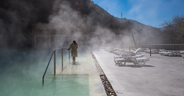 Person entering an outdoor thermal pool surrounded by steam, with mountains in the background and empty sun loungers by the pool
