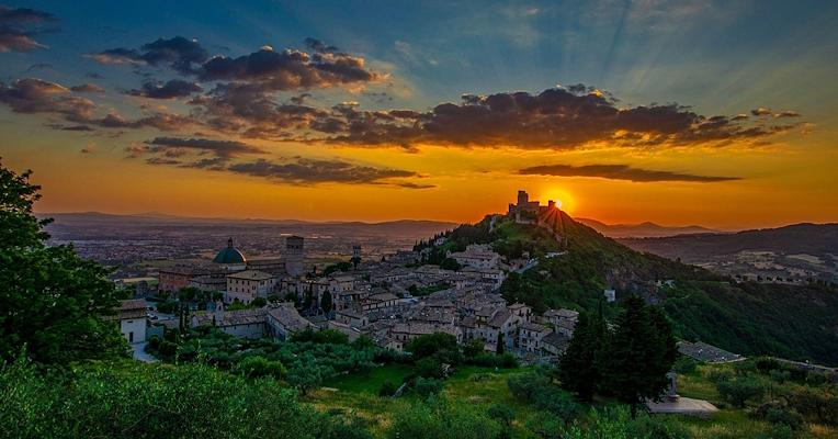 Panoramaansicht von Assisi bei Sonnenuntergang, mit der Sonne, die hinter einem Hügel untergeht, und einem Himmel in Orange- und Blautönen.