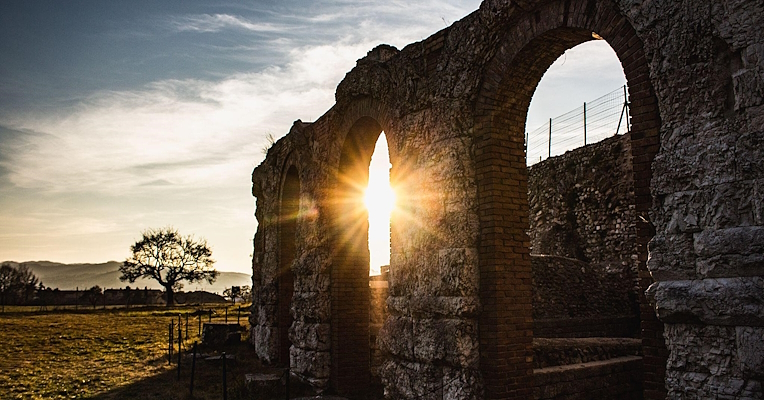 Sun setting behind an arch of the Roman Theatre of Gubbio, casting warm light and shadows over the surrounding landscape.