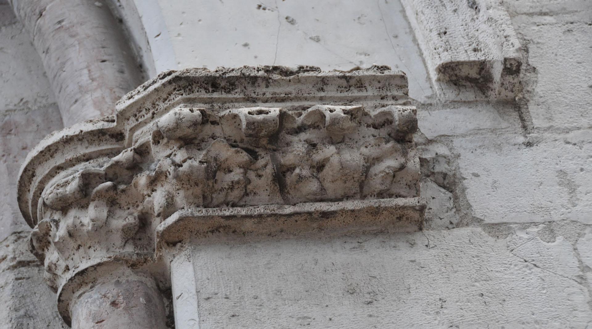 Detail eines steinernen, gemeißelten Kapitells mit vom Lauf der Zeit abgenutzten Ziermotiven an der Kirchenfassade.
