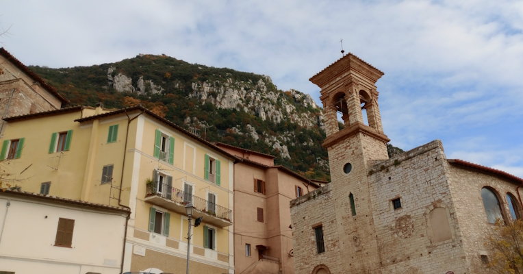 View of a stone bell tower and colorful houses with a mountain and a blue sky in the background.