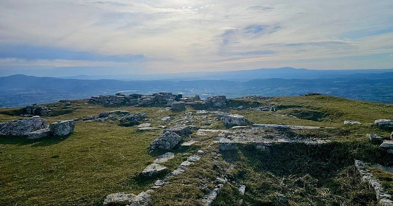Stone ruins on a grassy hilltop with a panoramic view of hills and mountains on the horizon under a cloudy sky.