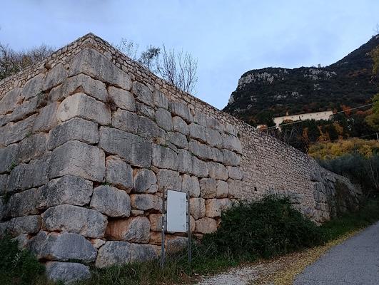 Alte Steinblockmauer entlang einer Bergstraße, mit Vegetation und Hügeln im Hintergrund.