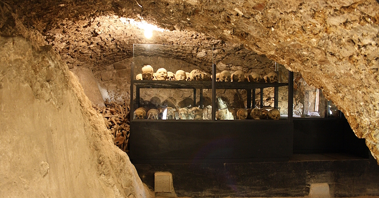 Underground crypt with human skulls and bones displayed in glass cases, lit by a single light bulb.