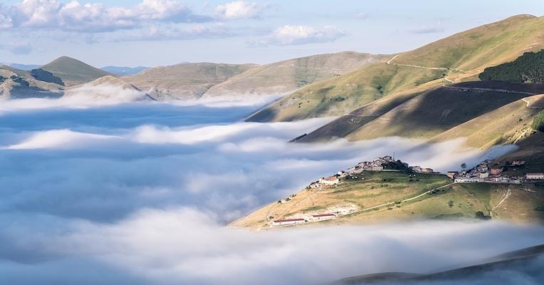 A village on a hill, surrounded by low-lying clouds amid green slopes and peaks bathed in the morning light.