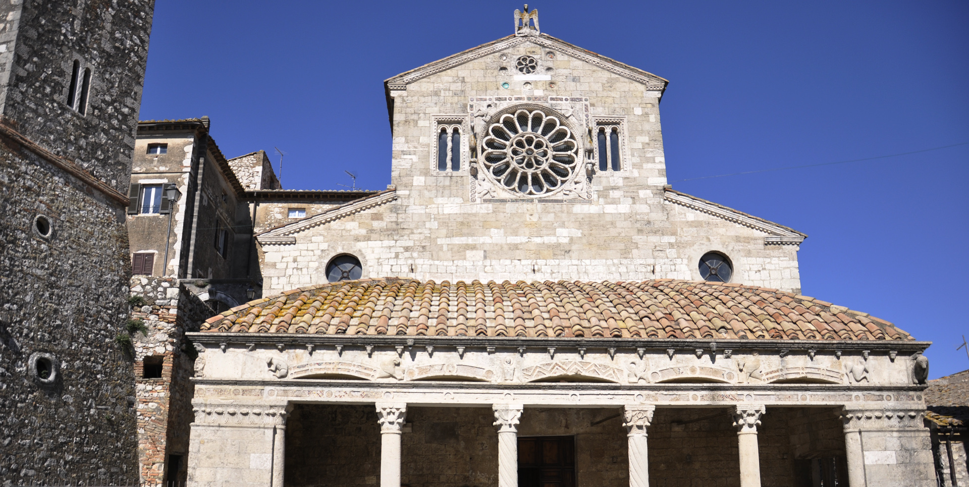 Steinfassade der Kirche Santa Maria Assunta mit zentraler Rosette und Säulenportikus unter blauem Himmel.