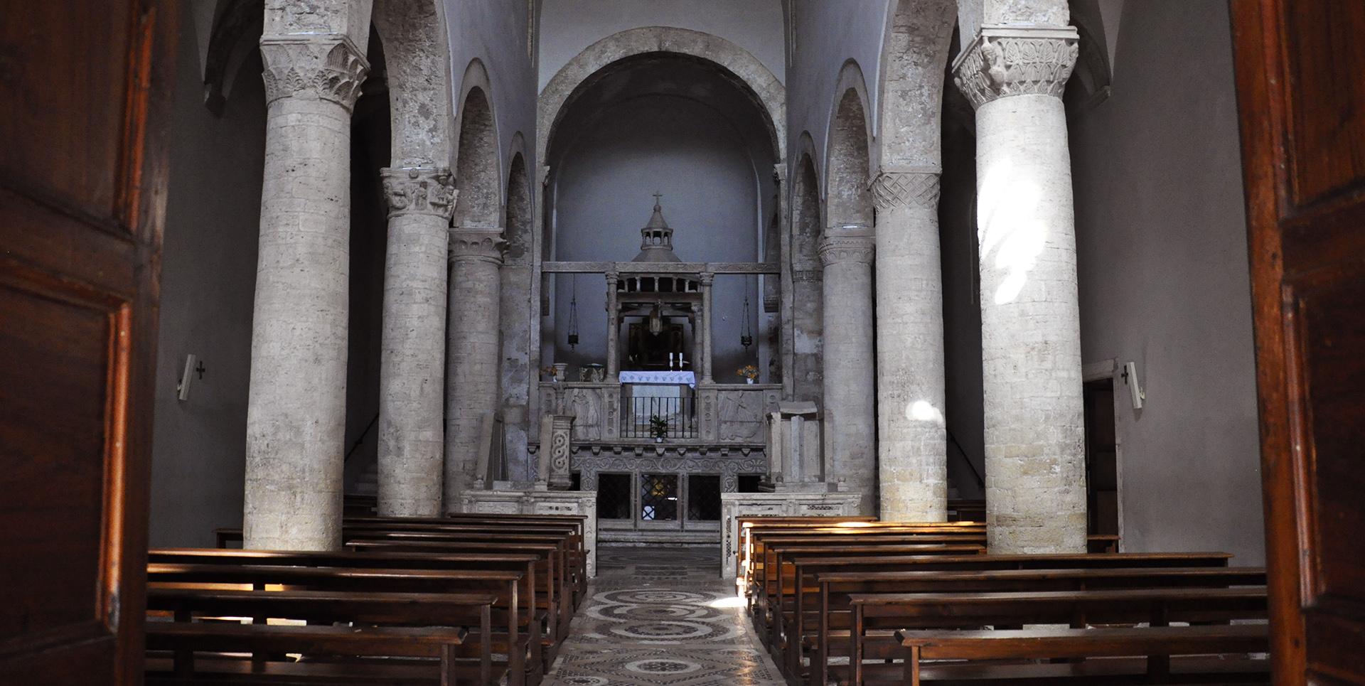 Interior and central nave of the Church of Santa Maria Assunta with marble columns, wooden pews, and raised altar at the back.