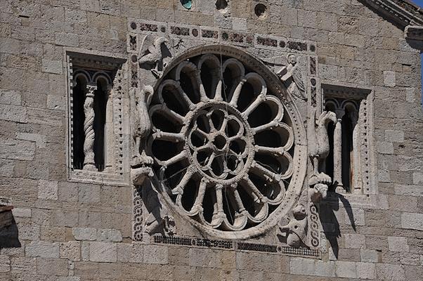 Romanesque-style rose window at the centre of the façade of the Church of Santa Maria Assunta, with sculpted decorations on the stone façade at the sides.
