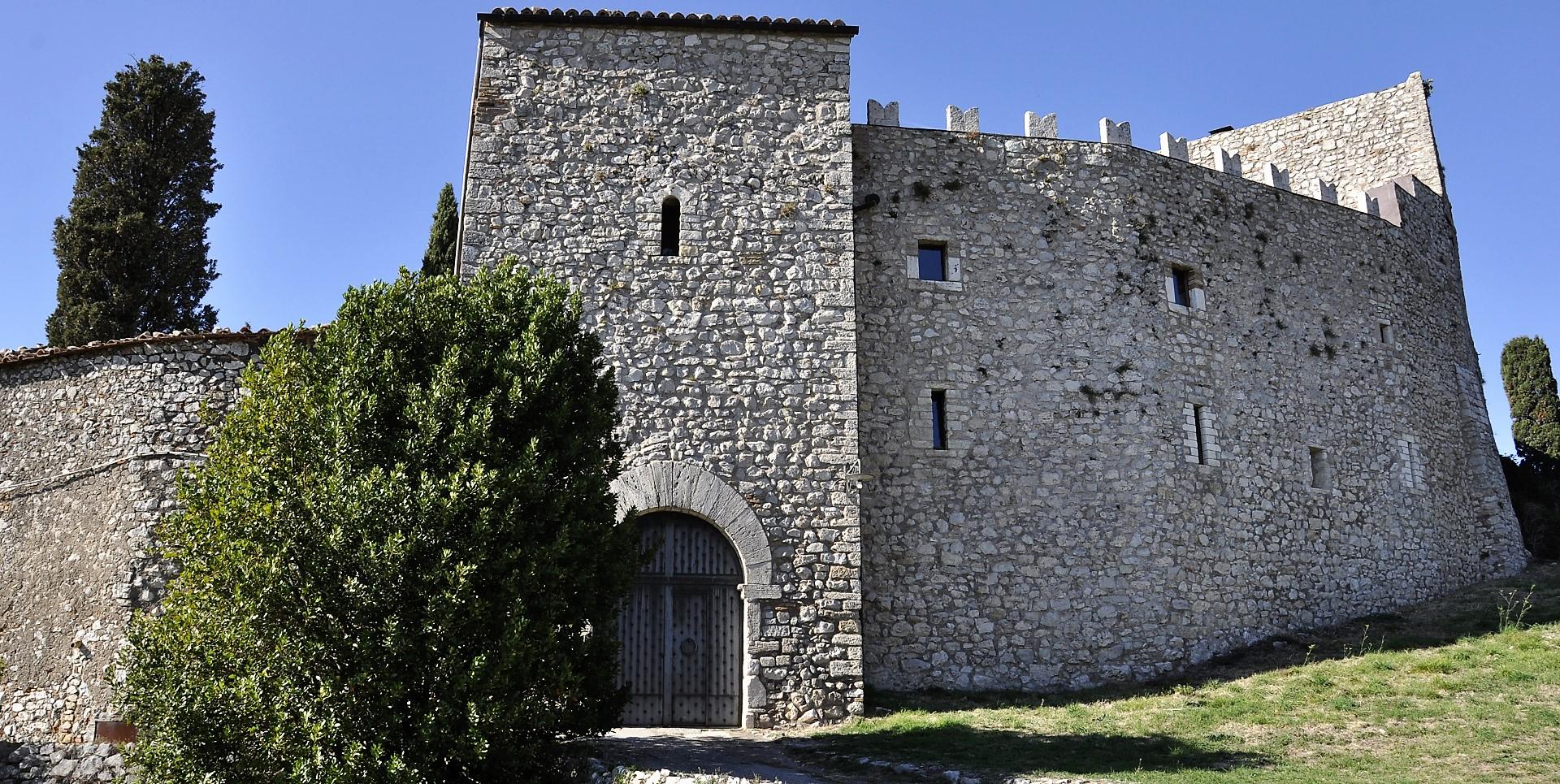 Stone façade of Castello del Poggio with a crenellated tower, arched doorway and fortified walls, nestled in greenery.