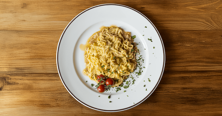 Tagliatelle with smoked tench, served with aromatic herbs and cherry tomatoes on a white plate, placed on a wooden table.