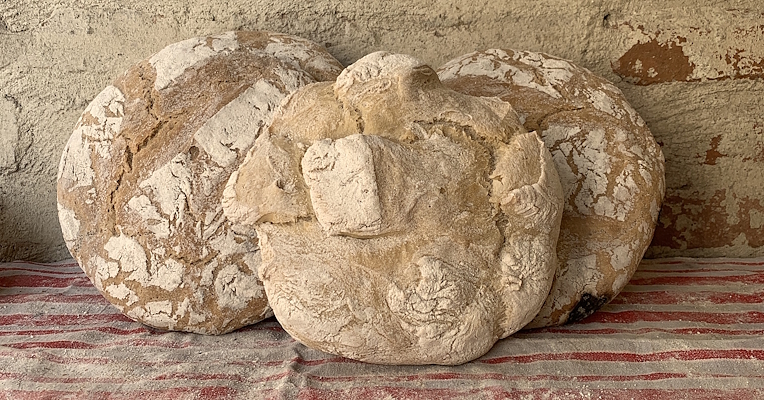 Three loaves of handmade unsalted bread dusted with flour on an ecru and red striped cloth, in front of a stone wall.