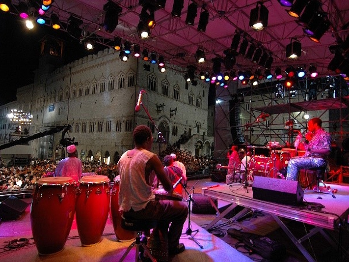 Blick von der Bühne eines Umbria-Jazz-Konzerts in Perugia, mit Musikern, die Schlaginstrumente vor dem Publikum spielen, beleuchtet von bunten Lichtern, mit dem Palazzo dei Priori im Hintergrund.