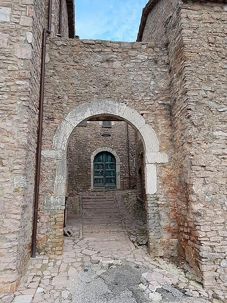 Steinbogen am Eingang der Burg von Macerino, von dem aus man eine Gasse mit einer Treppe und im Hintergrund eine grüne Tür erblicken kann
