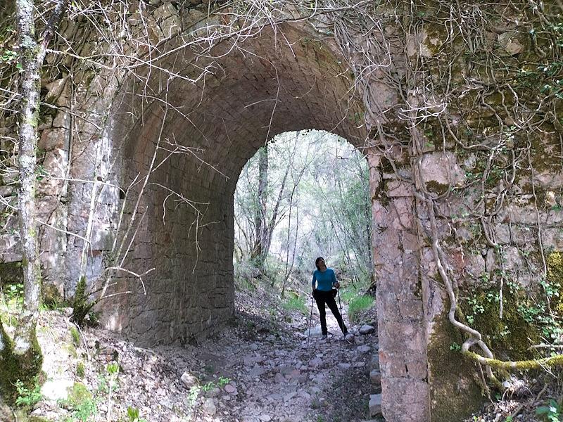 Wanderer unter einer alten Steinbrücke, bewachsen mit Moos und Kletterpflanzen, inmitten der Wälder der Monti Martani, in der Nähe von Macerino