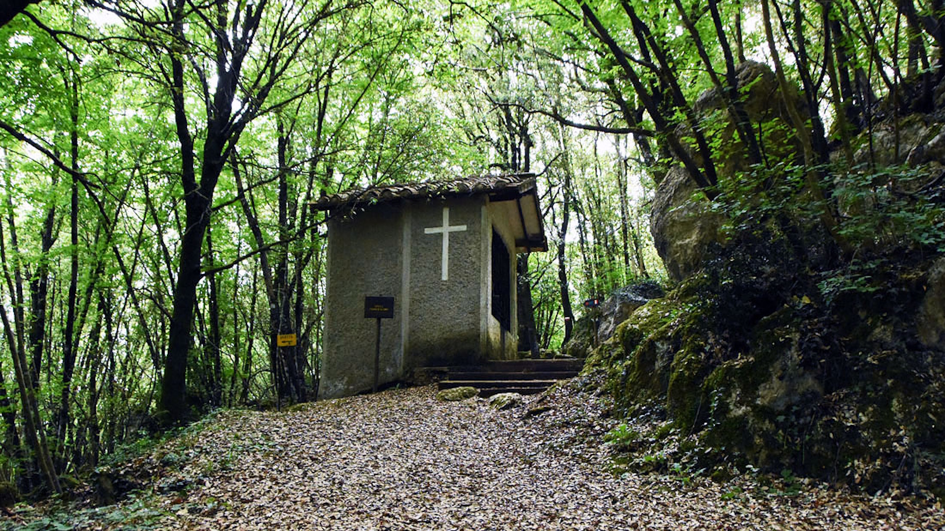 Small Franciscan chapel nestled in the woods, with a white cross on the side façade.