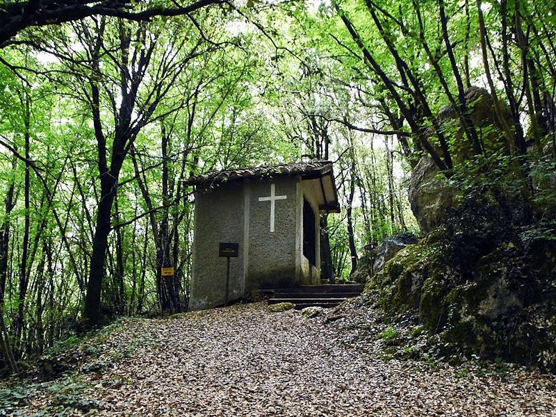 Kleine franziskanische Kapelle im Wald mit einem weißen Kreuz an der Seitenfassade.