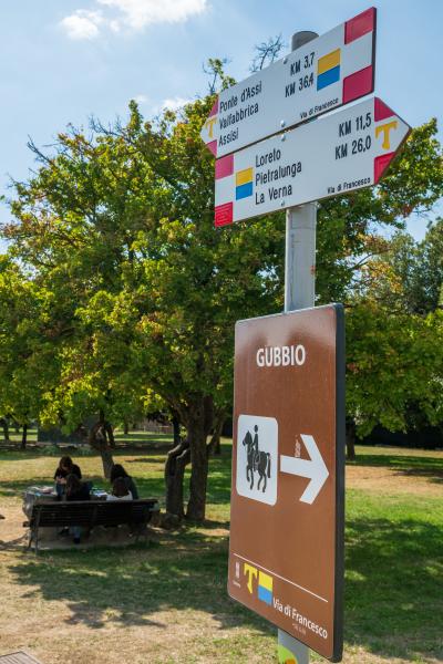 Placas da Via de Francisco em Gubbio com direções e distâncias para Assis e La Verna.