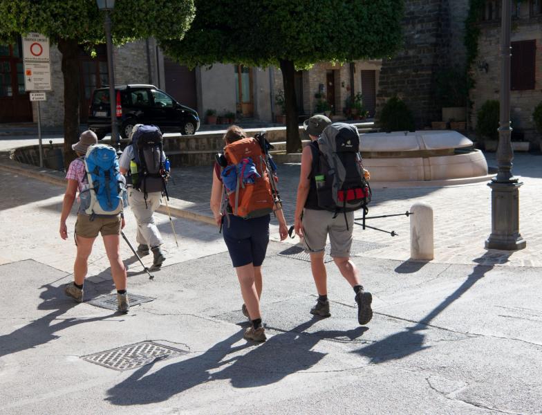 Grupo de peregrinos com mochilas e bastões de caminhada atravessa uma praça histórica da Via de Francisco.