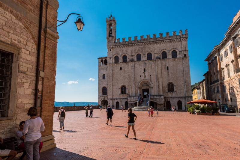 Piazza Grande a Gubbio con il Palazzo dei Consoli e persone che passeggiano nella piazza.