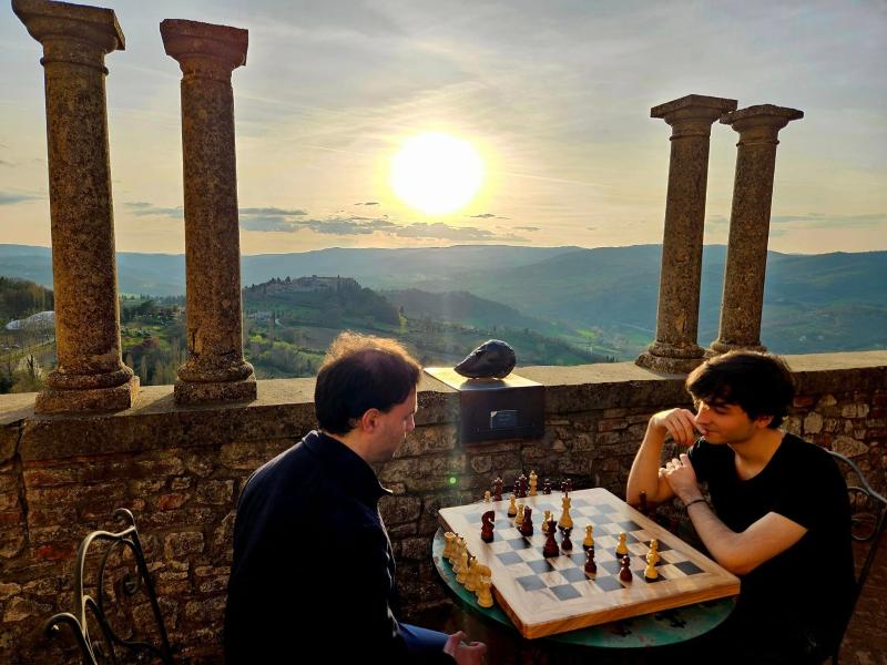 Two men play chess at sunset on a panoramic terrace with columns and Umbrian hills in the background.