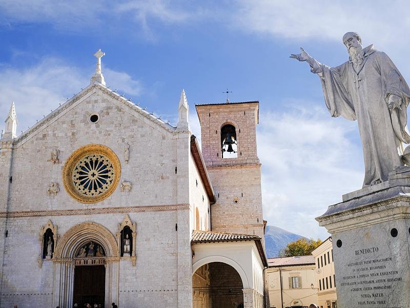 Statua di San Benedetto davanti alla basilica di Norcia, con facciata in pietra e cielo azzurro sullo sfondo